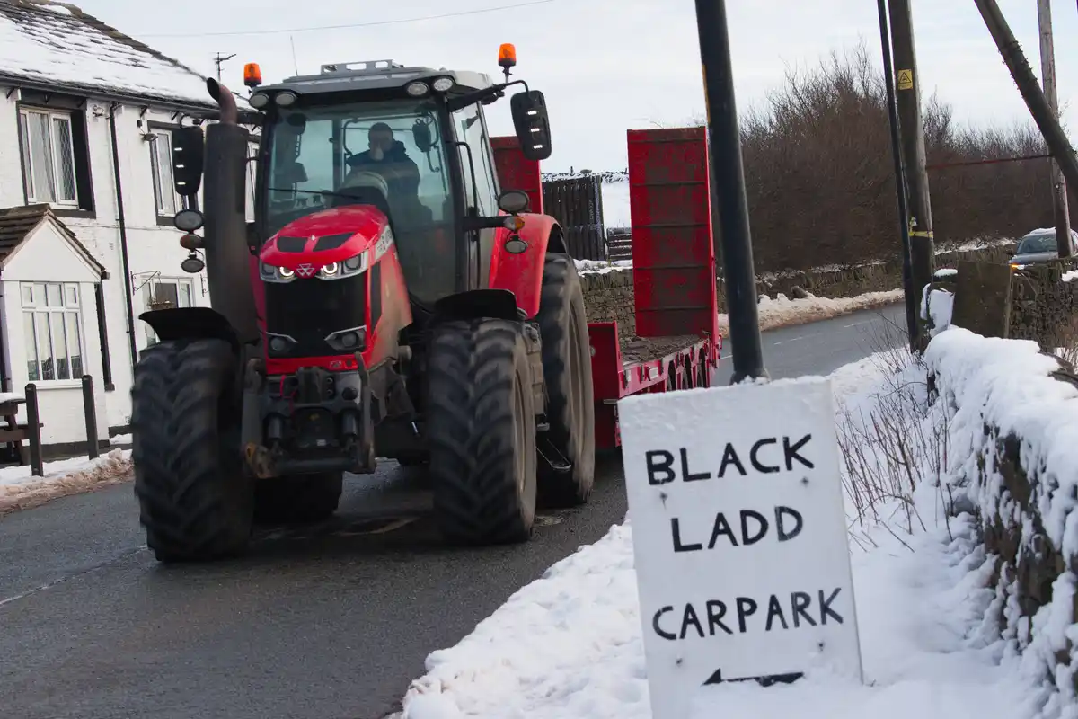 A red tractor with a large trailer is driving down a snowy rural road past a white sign that reads 'BLACK LADD CARPARK'. The…