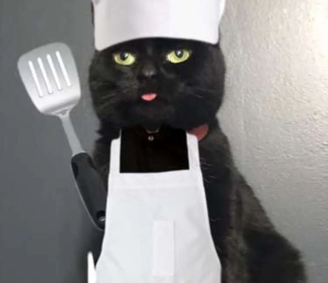 A black cat wearing a white chef's hat and apron, holding a white spatula in its paw, with its tongue slightly sticking out…