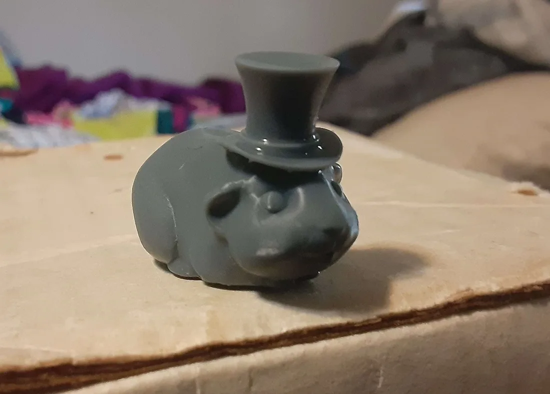 A small, gray, 3D-printed figurine of a sheep wearing a top hat sits on a cardboard surface. The sheep has a simple…