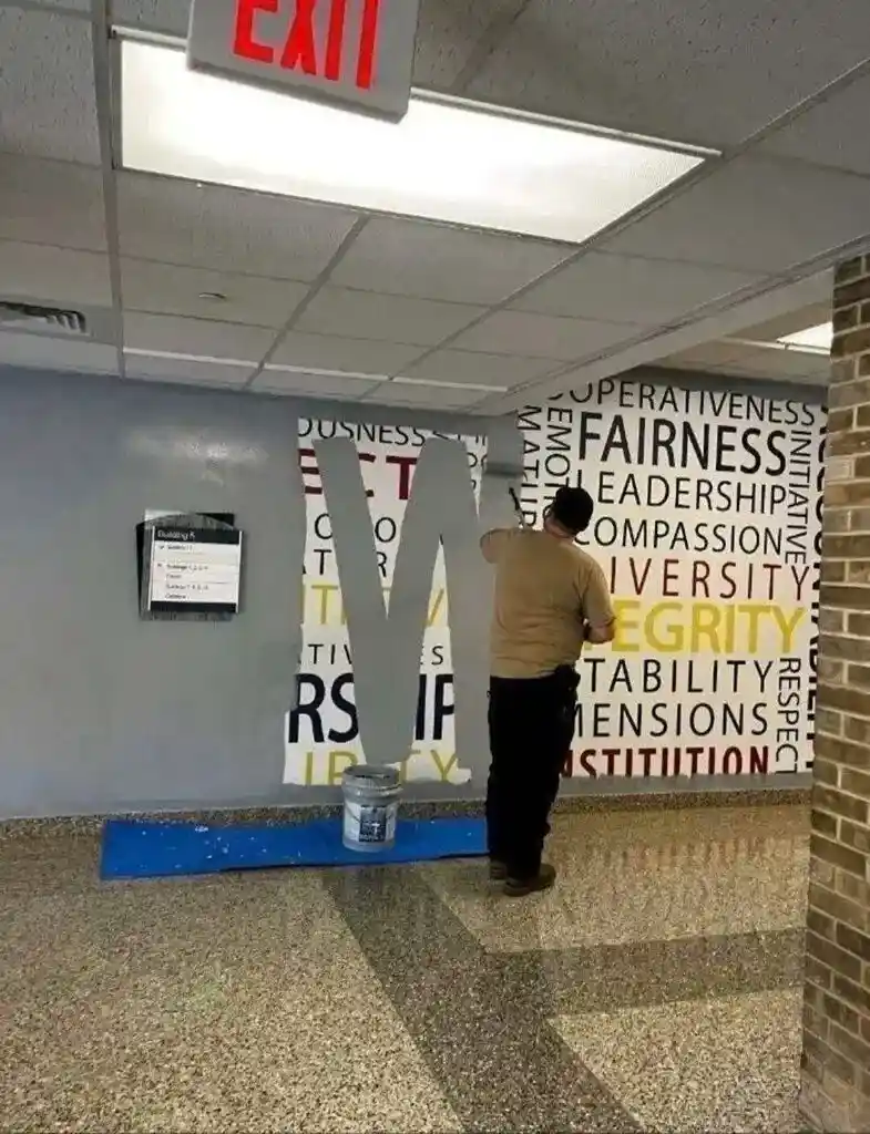 A person paints a large gray letter 'V' on a wall covered with words like 'fairness', 'leadership', 'compassion'…