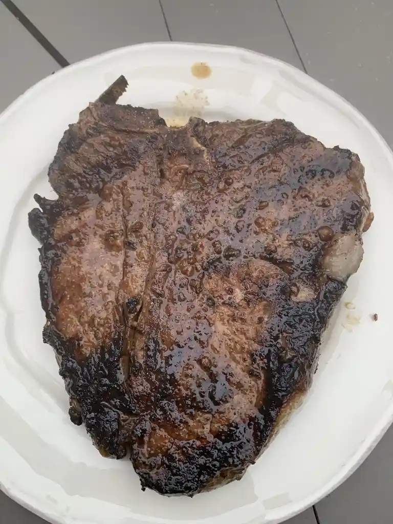 A close-up photo shows a large, heavily charred piece of meat on a white plate, with blackened edges and a glossy, burnt…