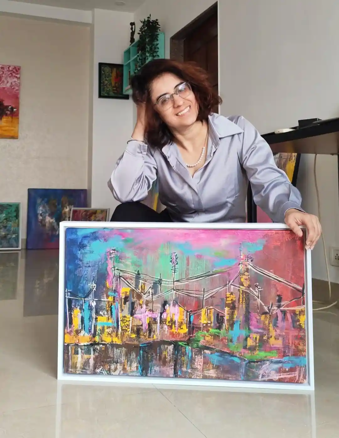 A woman with glasses and a light blue shirt smiles while holding a large, colorful abstract painting of a bridge. The…