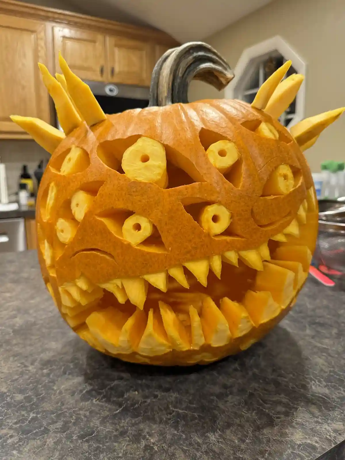 A highly detailed, intricately carved Halloween pumpkin sits on a dark countertop in a kitchen, featuring pineapple slices…