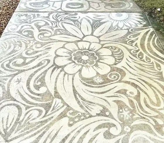 A close-up photo of a concrete sidewalk decorated with intricate, swirling white floral and leaf patterns painted on its…