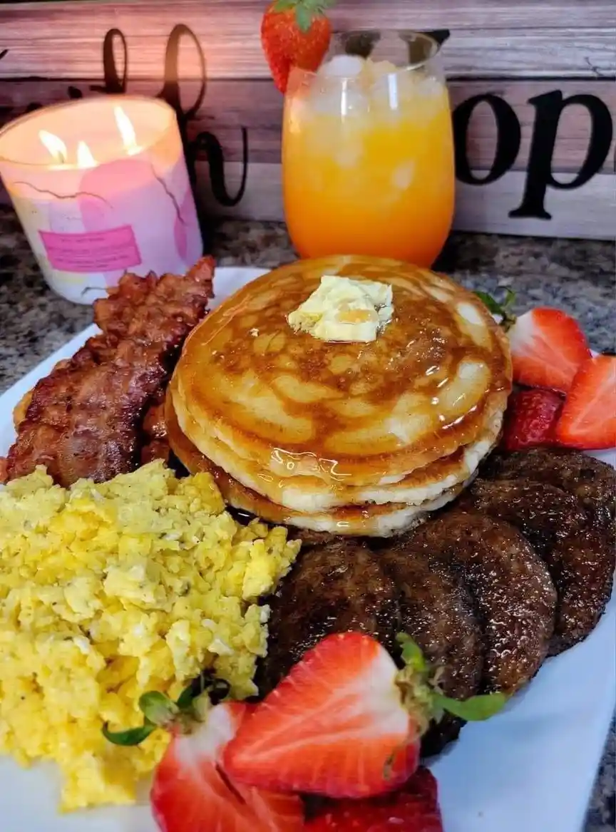 A plate of breakfast food including pancakes with syrup and butter, scrambled eggs, bacon, sausage patties, and sliced…