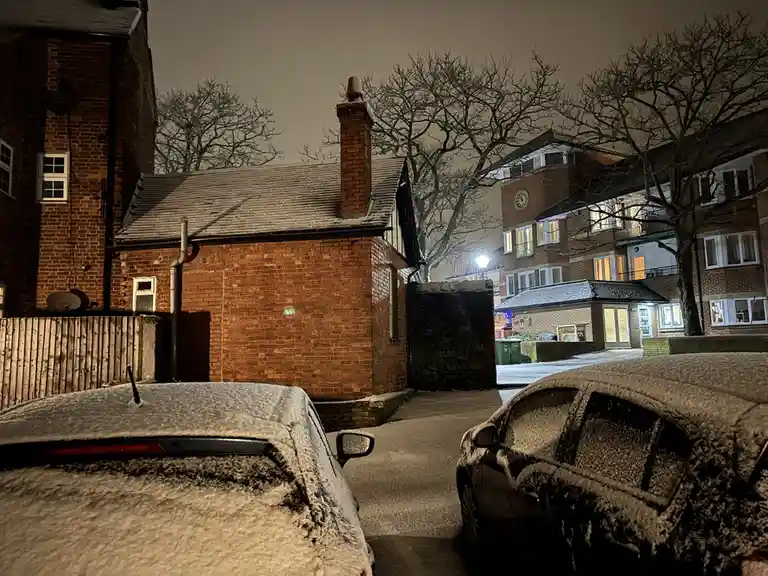 A nighttime street scene covered in snow, showing two cars parked on either side of a narrow alleyway, both heavily…