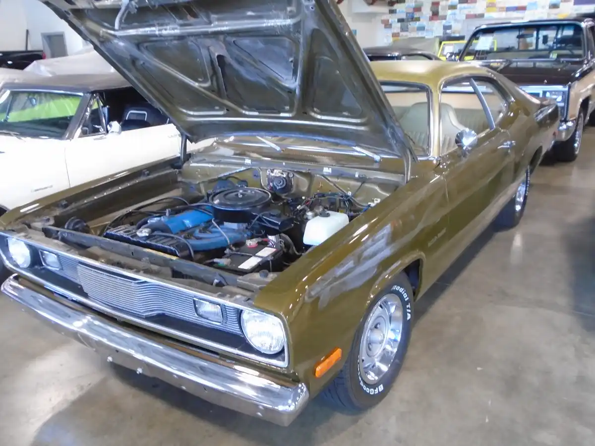 A vintage olive-green muscle car with its hood open, revealing a blue engine, is parked indoors among other classic cars…