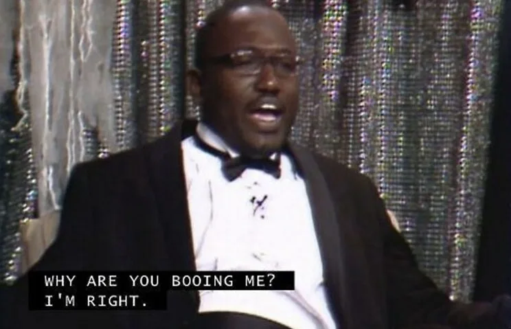 A man in a tuxedo with glasses and a bowtie is shown mid-speech, looking confused and slightly annoyed, with a sparkly…