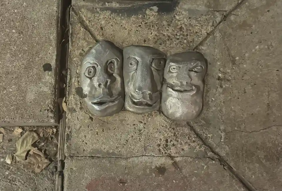 Three metallic, crudely sculpted faces with exaggerated, unsettling expressions are embedded in a concrete sidewalk. The…