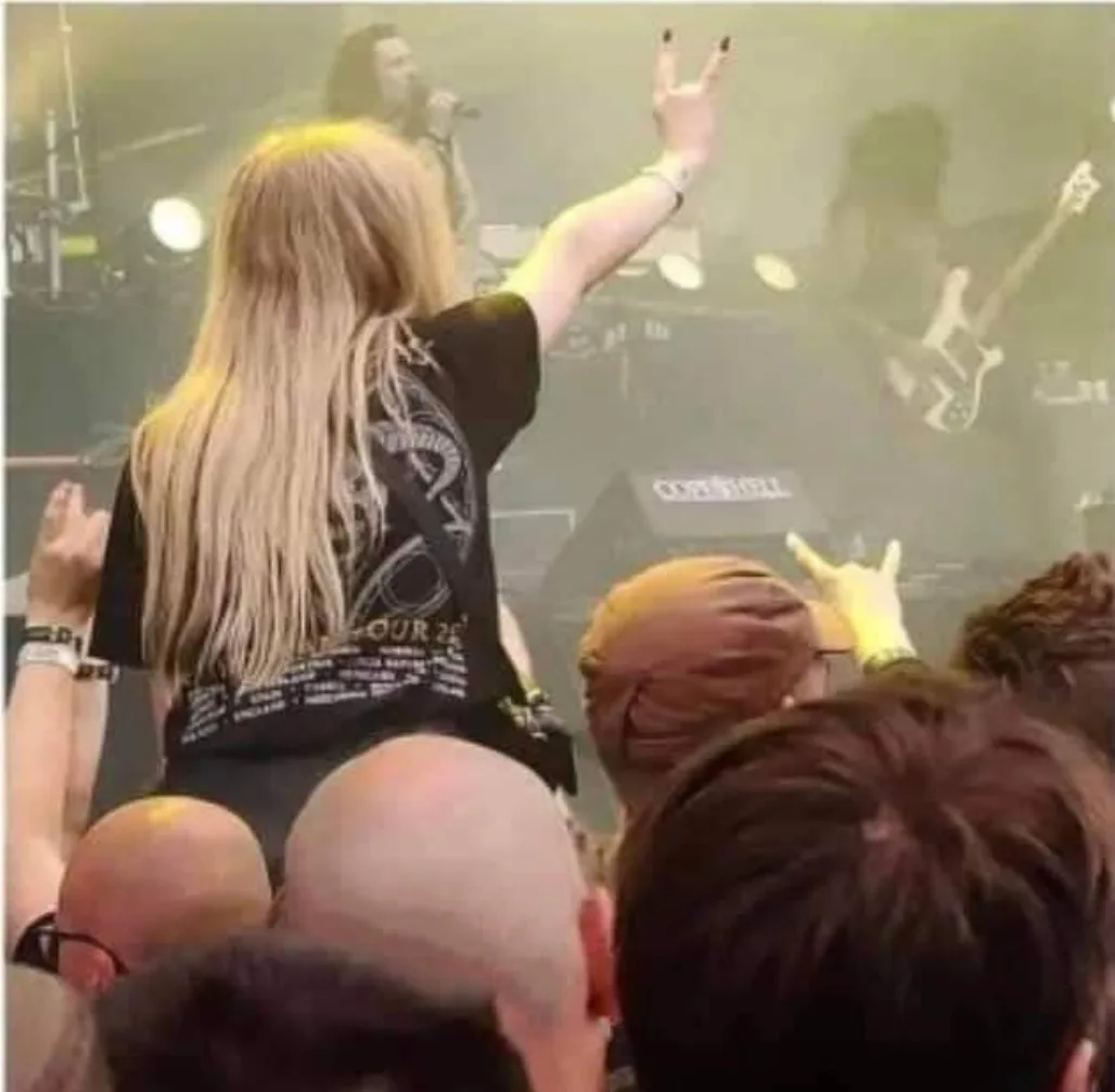 A person with long blonde hair, seen from behind, is at a concert, wearing a black t-shirt with a graphic and text, and…