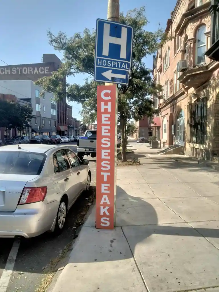 A street scene shows a blue hospital sign with an arrow pointing left, mounted on a pole that also has a vertical red sign…