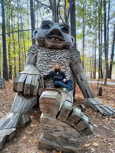 A man with long hair and glasses sits casually on the knee of a giant, stylized wooden sculpture of a creature resembling a…