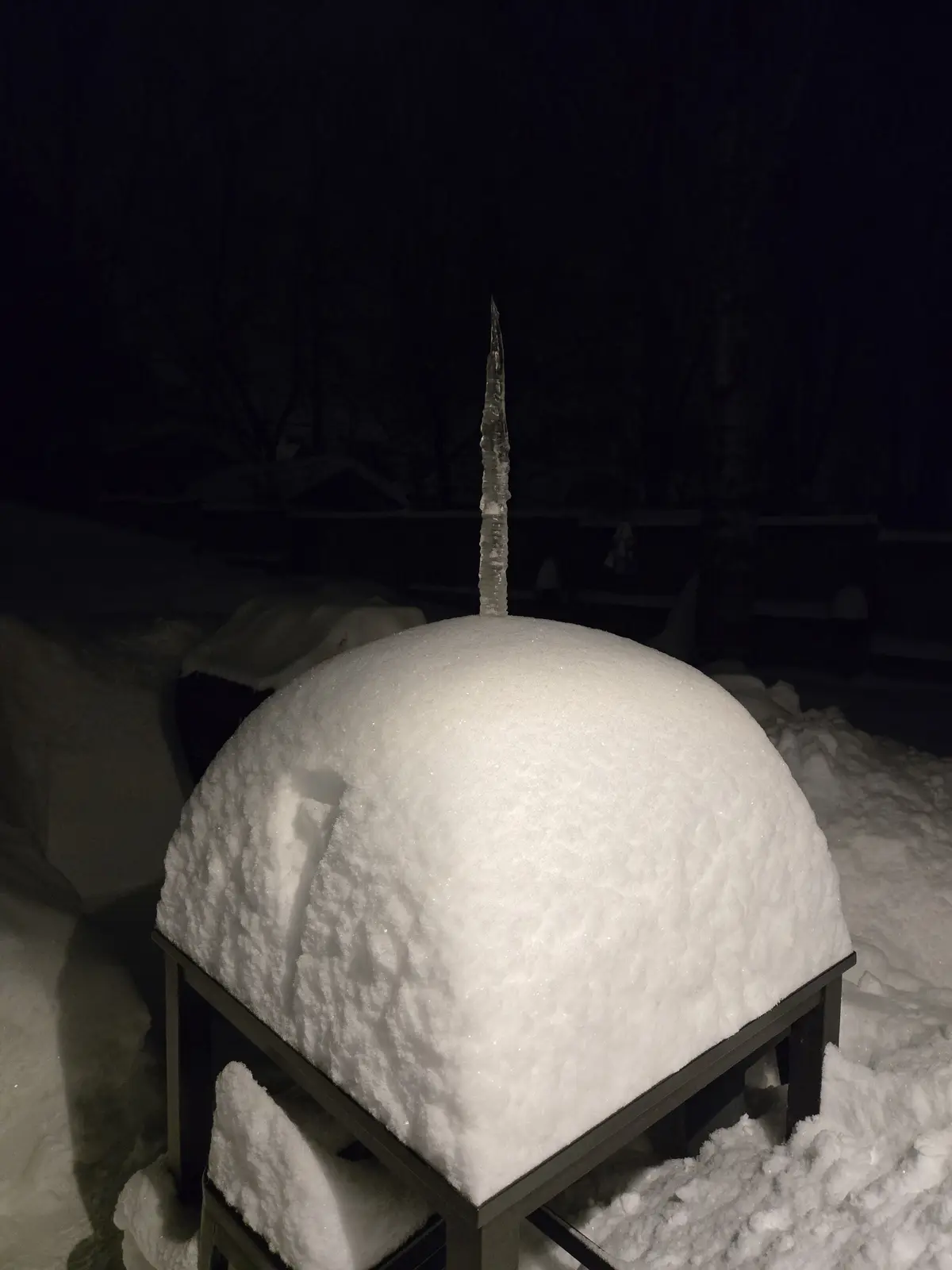 A snow dome, resembling an igloo, sits atop a black metal frame in a snowy yard at night. A long, clear icicle protrudes…