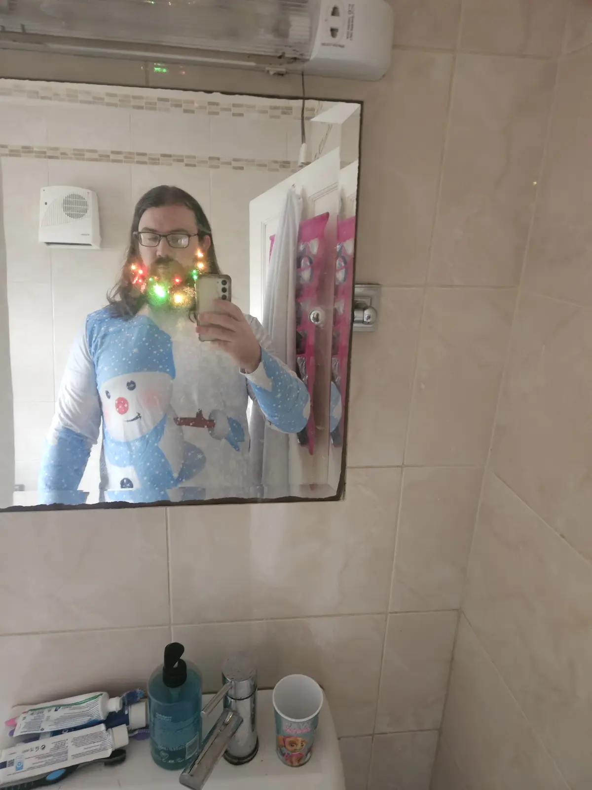 A person with long hair and glasses takes a mirror selfie in a bathroom while wearing a snowman-themed pajama top. They hold…