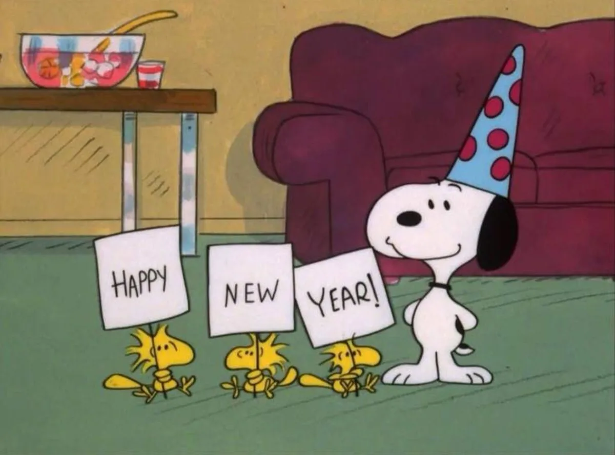 In a cartoon scene, Snoopy wears a blue party hat and stands beside three small yellow birds holding individual signs that…