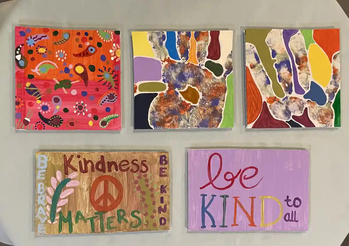 Five handmade signs are displayed on a table. Three colorful abstract paintings are at the top, and two wooden signs with…