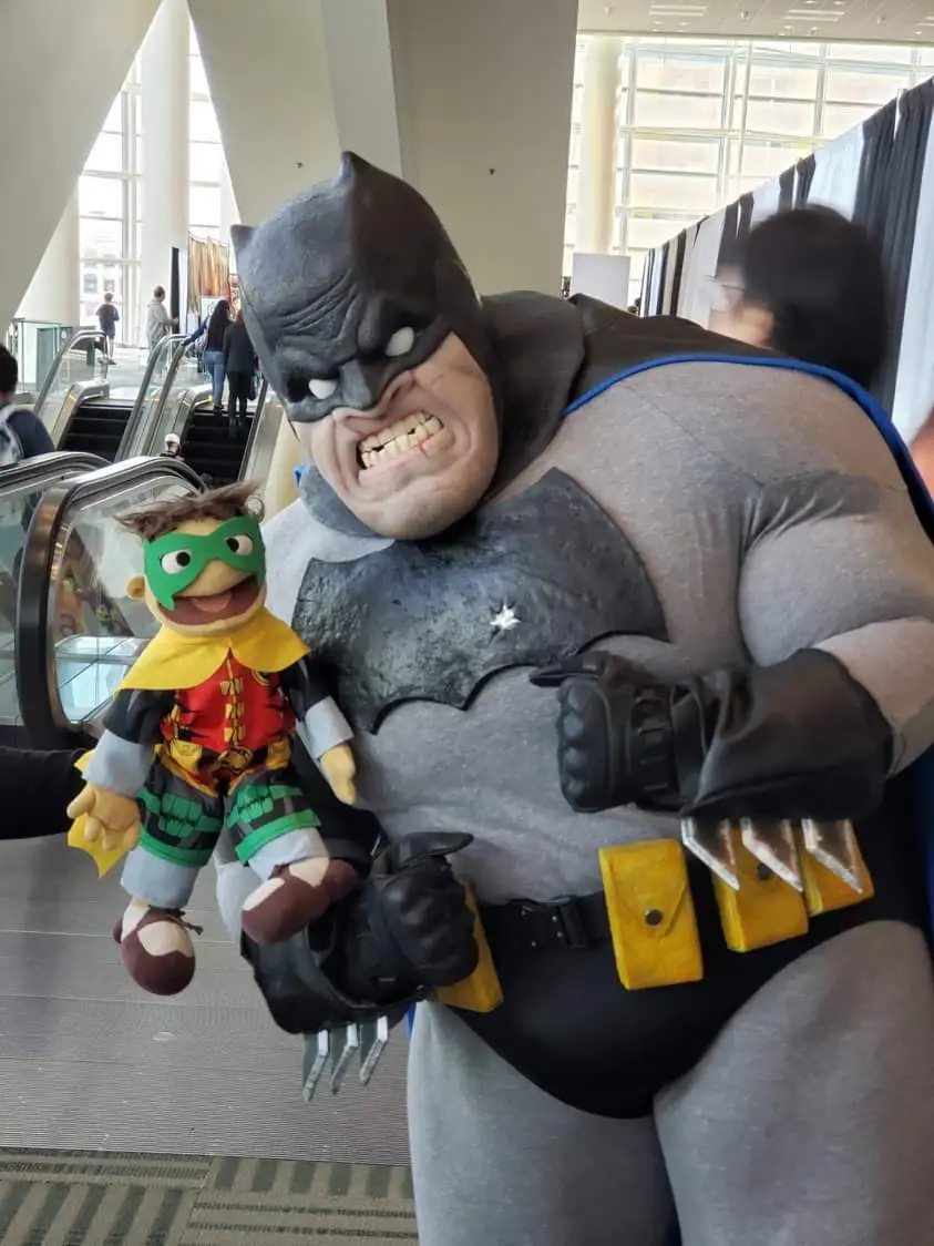 A person in a bulky, grey Batman costume with a large, angry mask and clawed gauntlets is holding a small, smiling Robin…