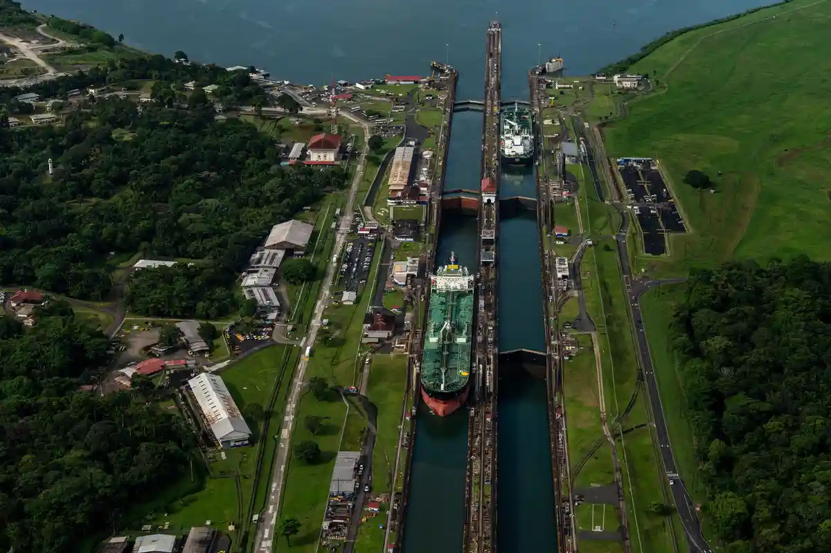 An aerial view of the Panama Canal with two large cargo ships passing through the locks, surrounded by green hills and…