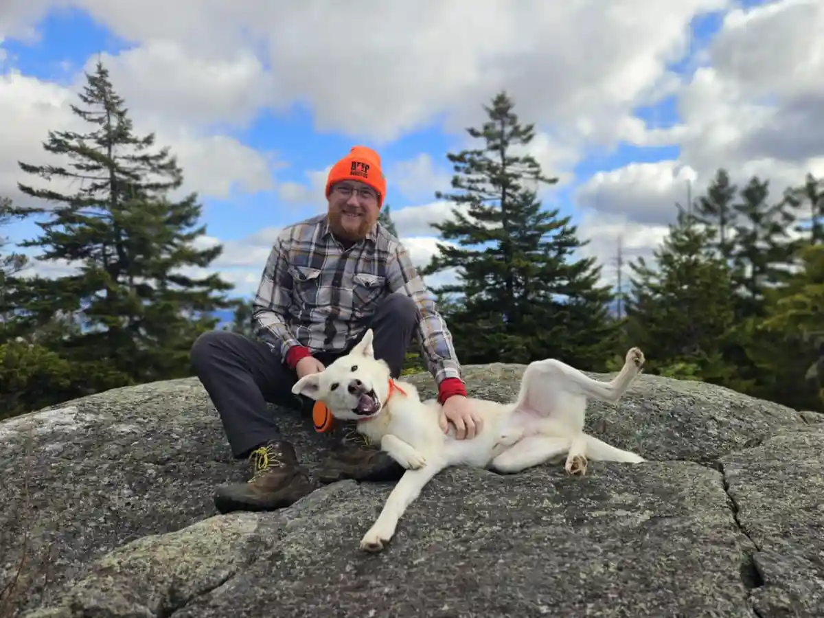 A man with a beard and glasses wearing an orange beanie and plaid shirt sits on a large gray rock outdoors with a white dog…