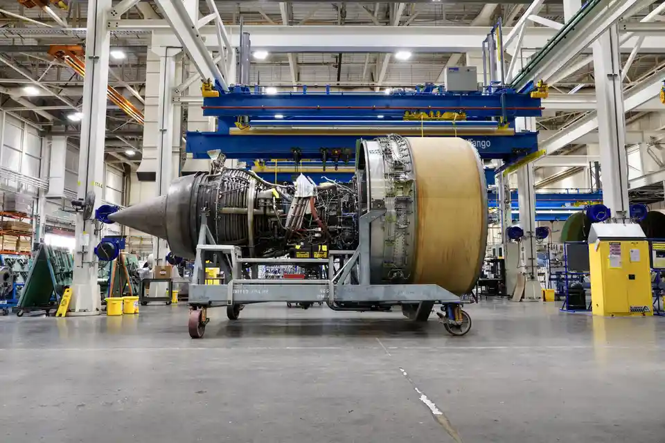 A large jet engine is shown on a maintenance cart inside a spacious industrial hangar, surrounded by equipment and overhead…