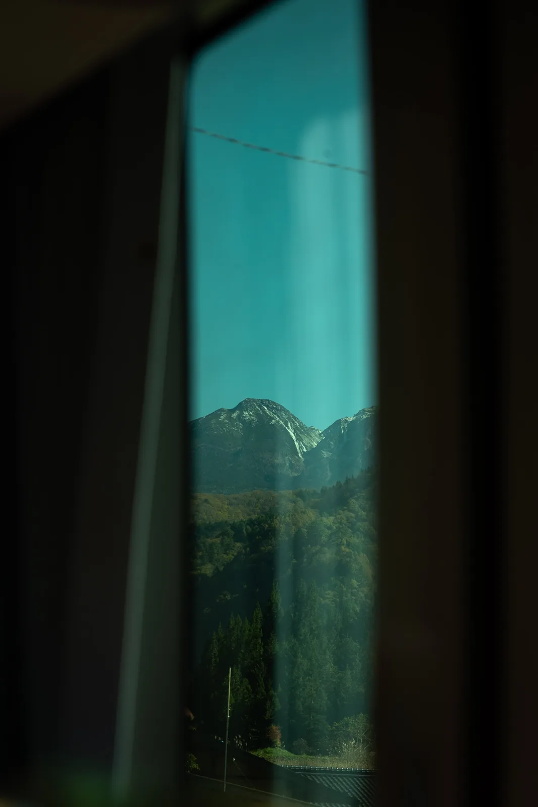 A view through a window frame shows a mountain range under a teal sky, with forests below and a thin wire across the top…