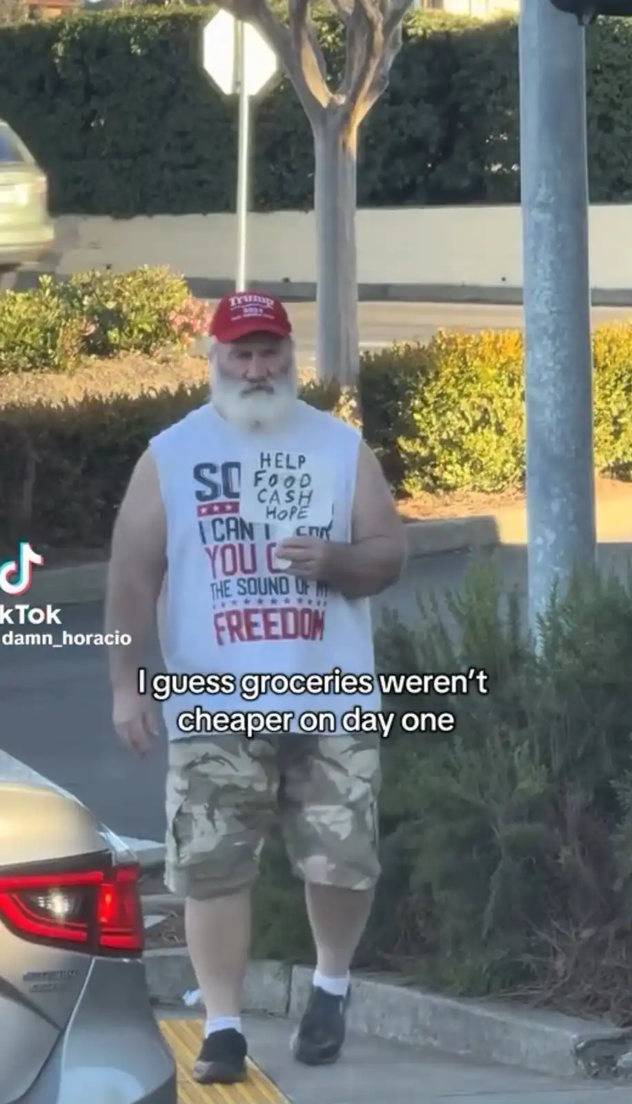 A man with a long white beard and a red 'Trump 2024' cap walks on a sidewalk near a parked car, wearing a white tank top…