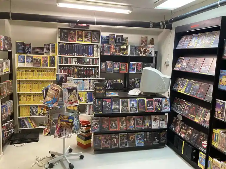A photo of a cluttered, retro video store filled with shelves of VHS tapes, video game consoles, and magazines, featuring a…