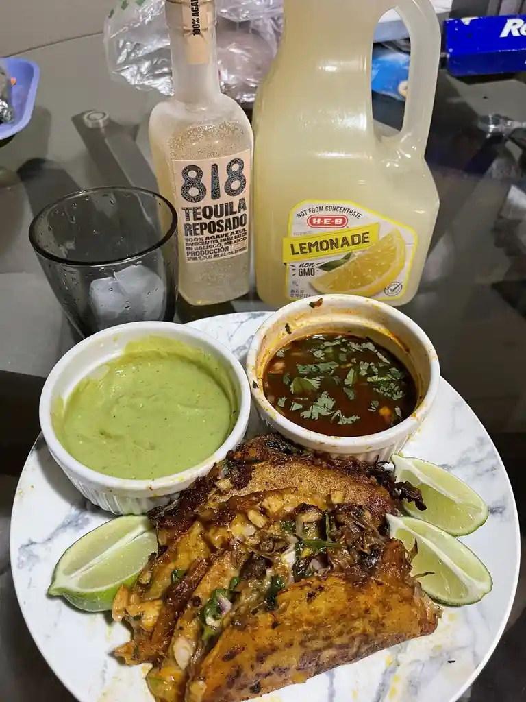 A plate of tacos with two small bowls of sauce and lime wedges sits on a table, next to a bottle of 818 Tequila Reposado and…