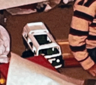 A low-resolution image shows a child's toy ambulance, white with black detailing, lying on its side on a red surface. A…