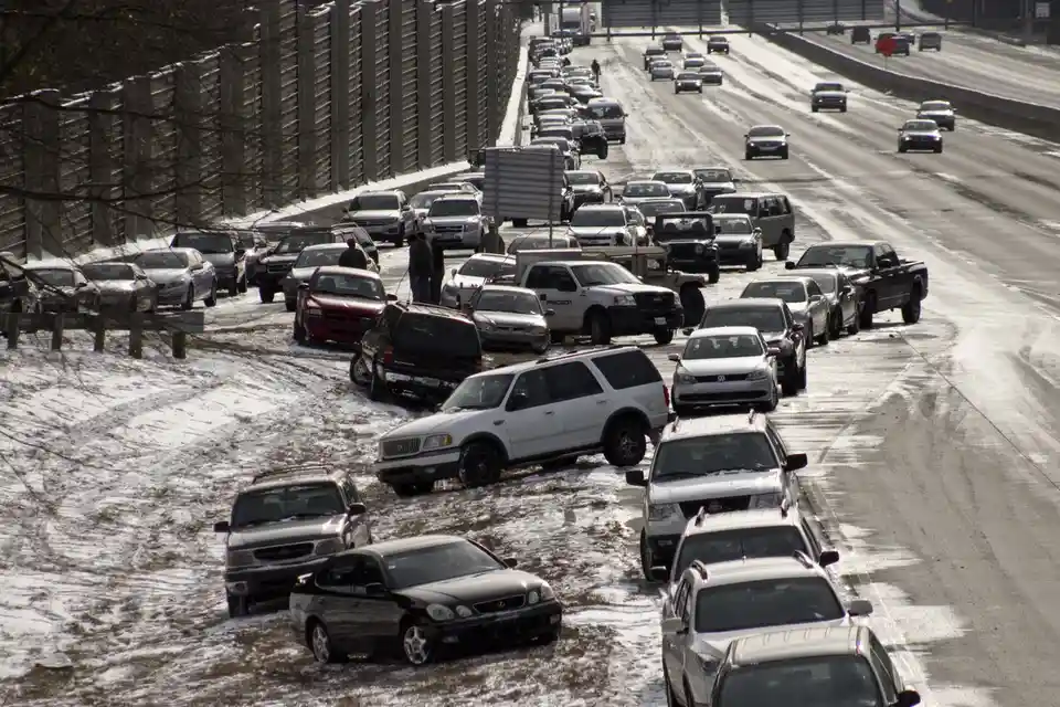 A snowy highway scene shows numerous cars abandoned or stuck off the road in a snowbank, while traffic continues to flow…