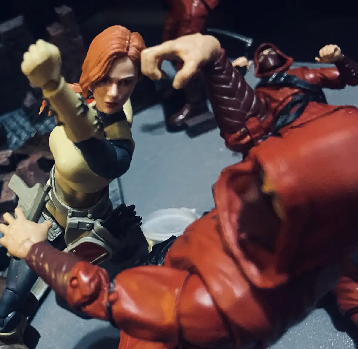 A close-up photo of action figures depicts a female character with red hair in a combat stance, facing down a larger male…