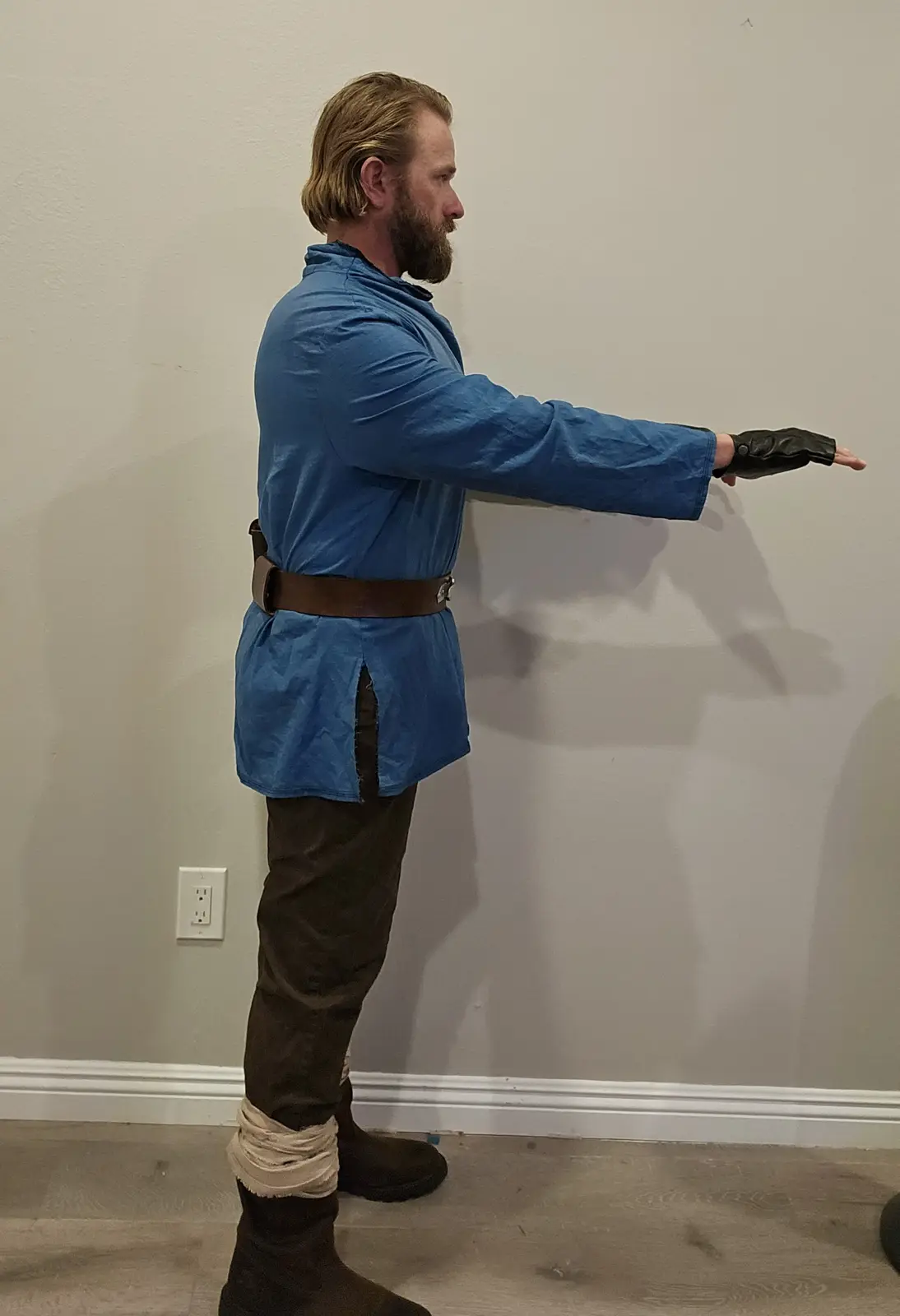 A man in a blue Jedi robe and brown pants, with a black fingerless glove, stands in profile against a plain wall, extending…