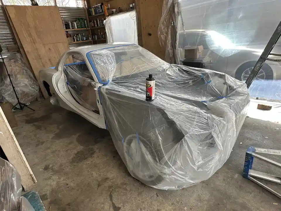 A white car body in a garage is mostly covered in clear plastic wrap, secured with blue painter's tape. A can of automotive…