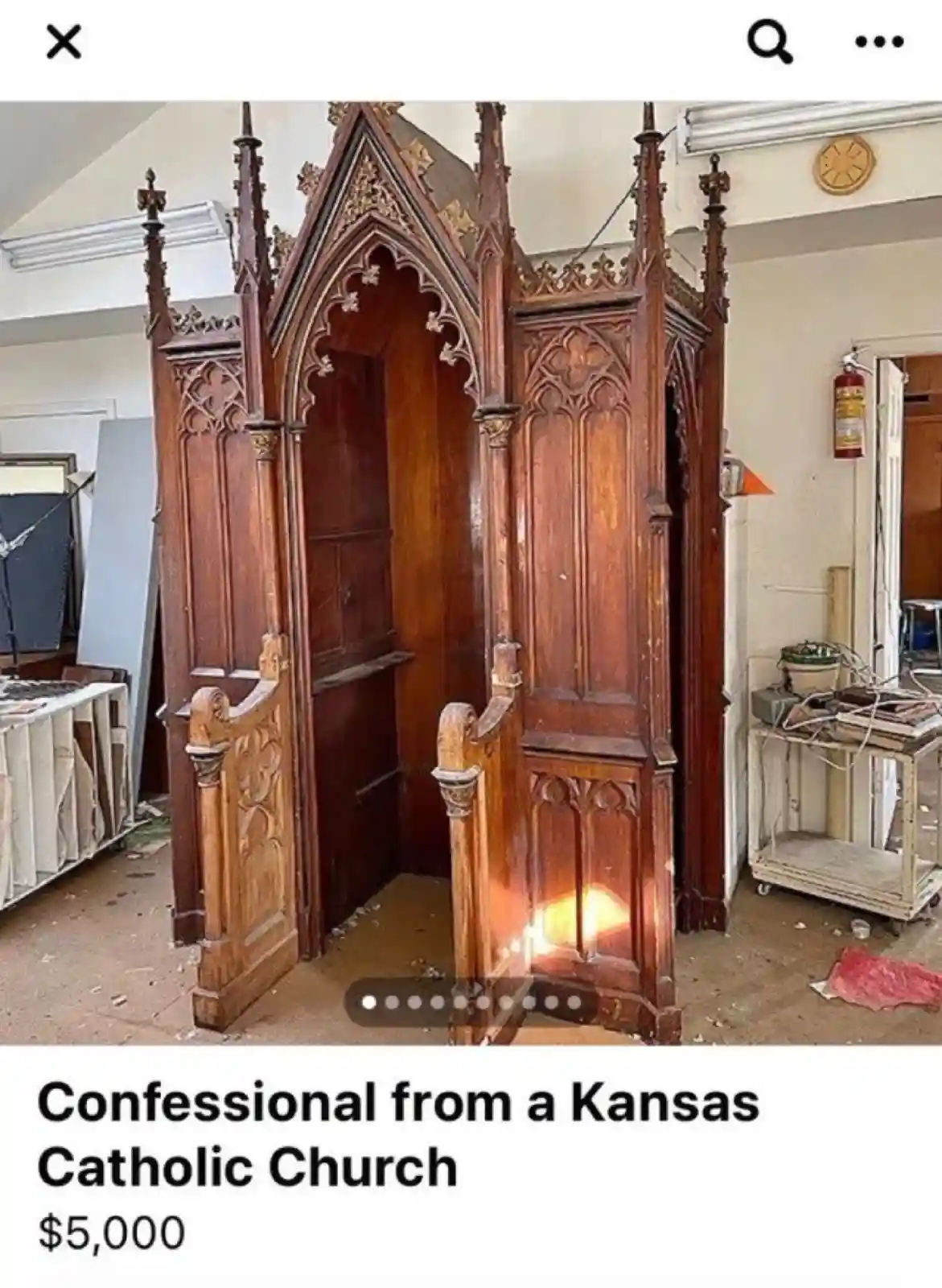 An ornate, wooden, Gothic-style confessional booth stands in a cluttered, unfinished room with debris on the floor and a…