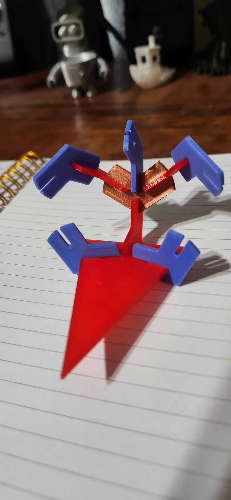 A 3d printed model of a character from a video game sits on a lined notebook, featuring a red triangular base, red arms, and…