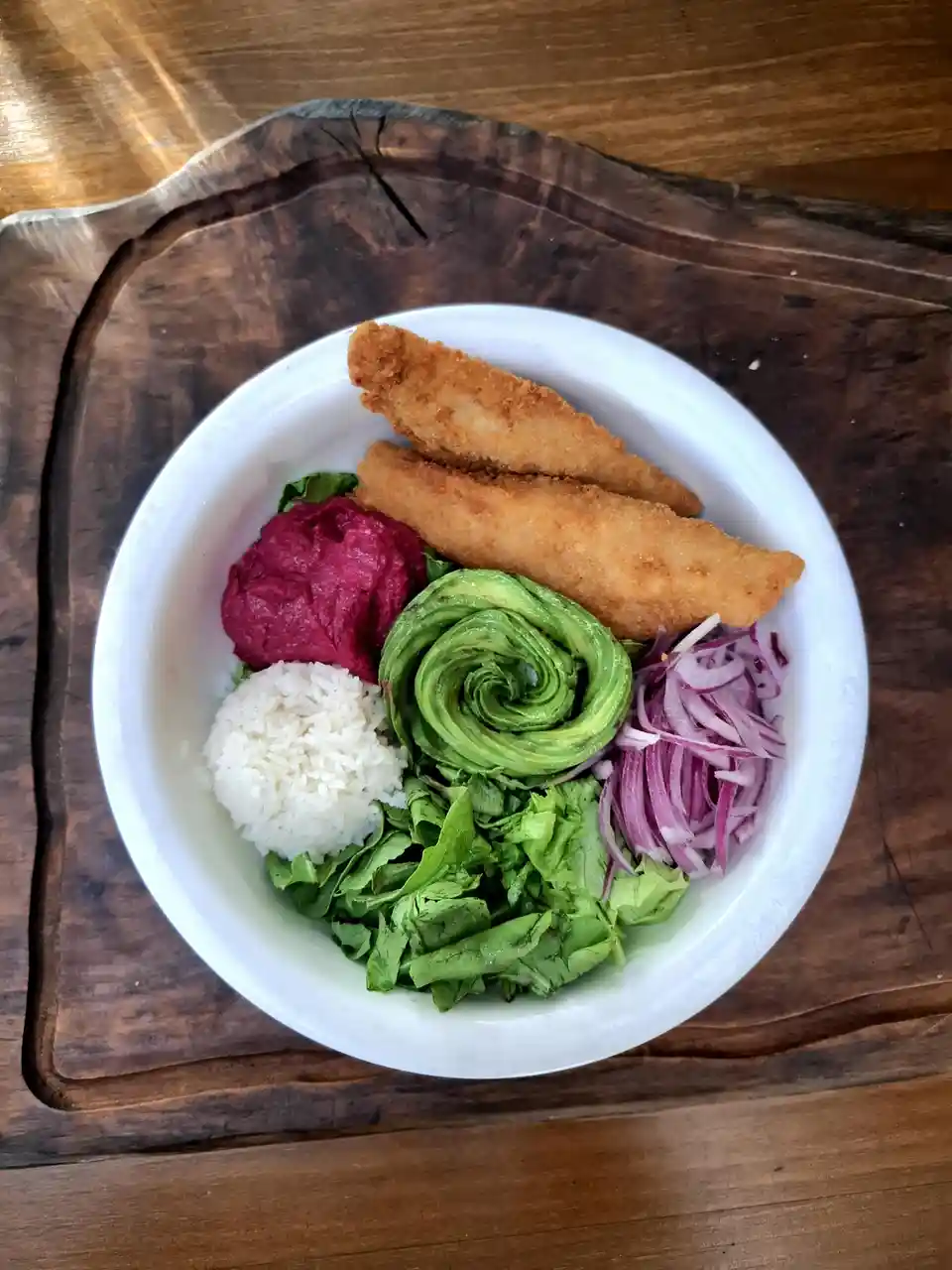 A top-down photo of a white bowl filled with colorful, neatly arranged food items including fried fish sticks, a pink beet…