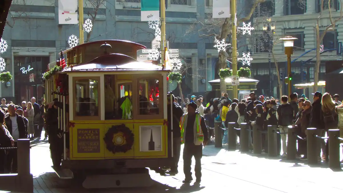 A yellow San Francisco cable car decorated with Christmas wreaths and snowflake lights is stopped at a crowded street…