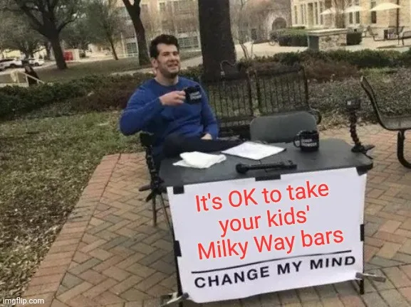 A man in a blue sweatshirt sits at a small outdoor table with a sign that reads 'It's OK to take your kids' Milky Way bars…