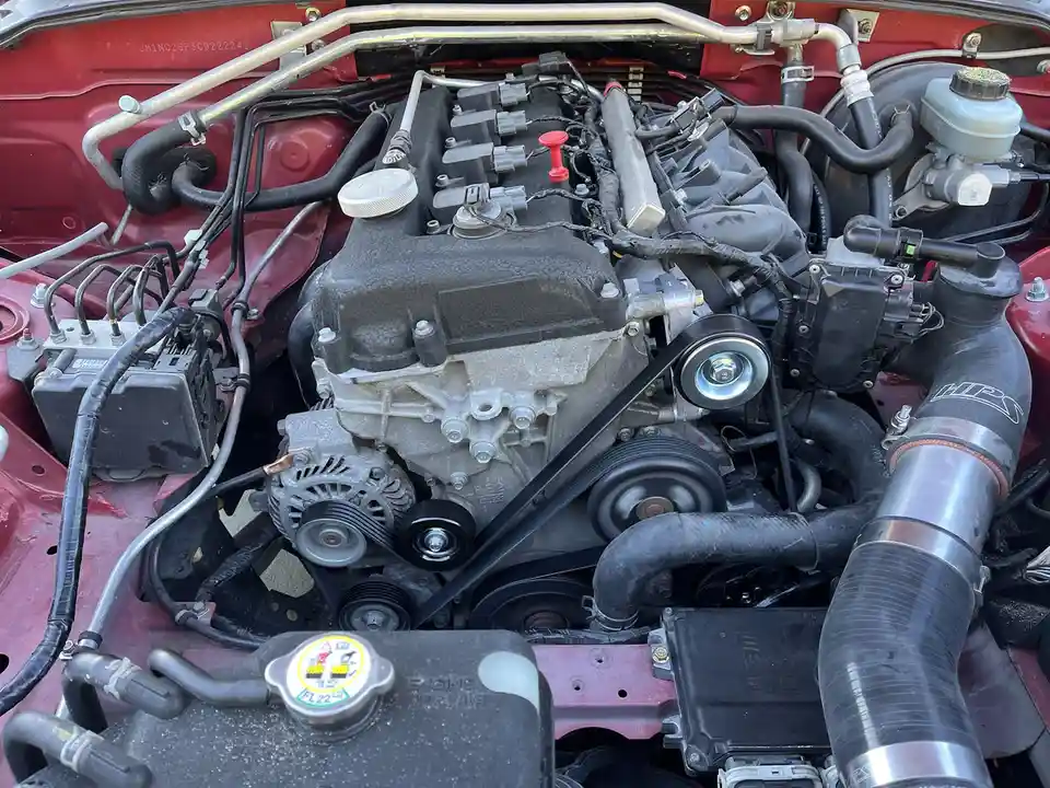 A close-up photo of a red car's engine bay showing a complex engine with various hoses, belts, and components like an…