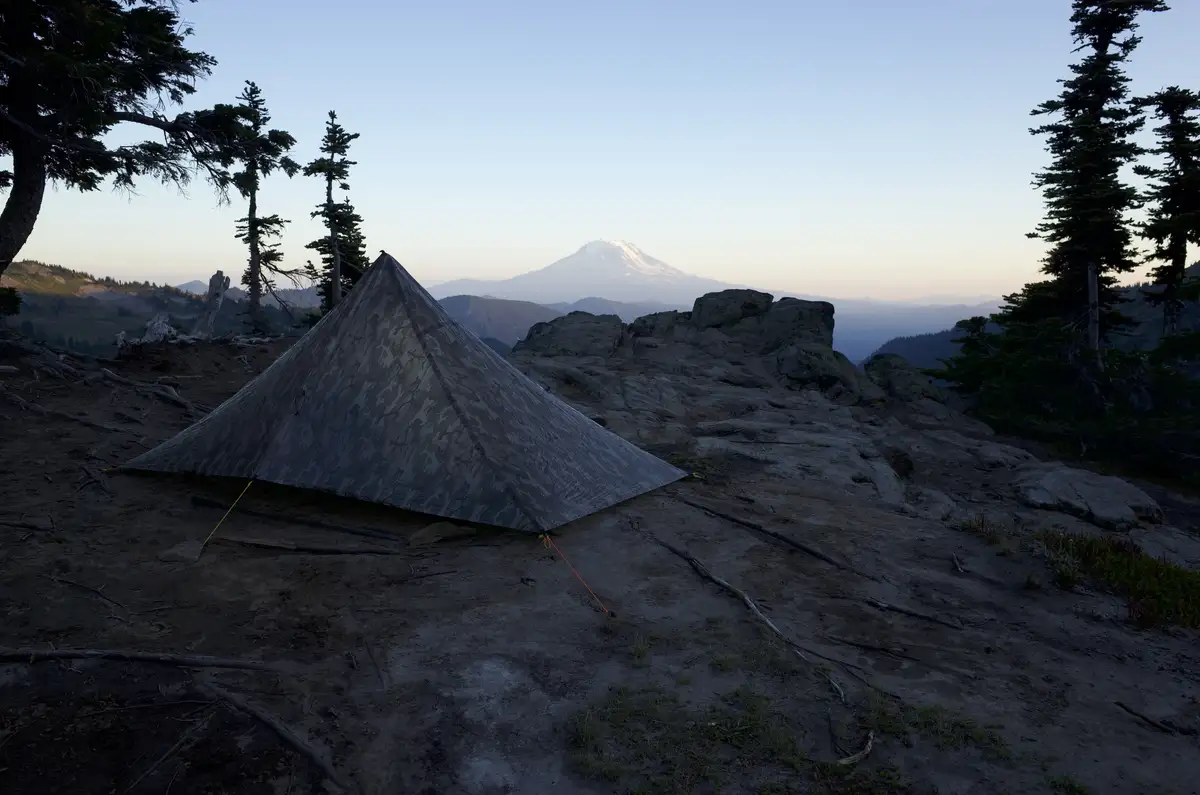 A camouflage-patterned, pyramid-shaped tent is pitched on a rocky, tree-dotted hillside at dawn or dusk, with a majestic…