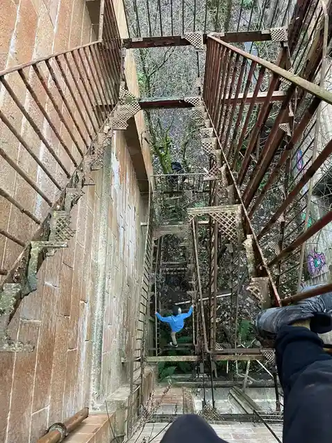 A first-person perspective photo shows a person's gloved hand and leg at the top of a rusty, dilapidated metal staircase…