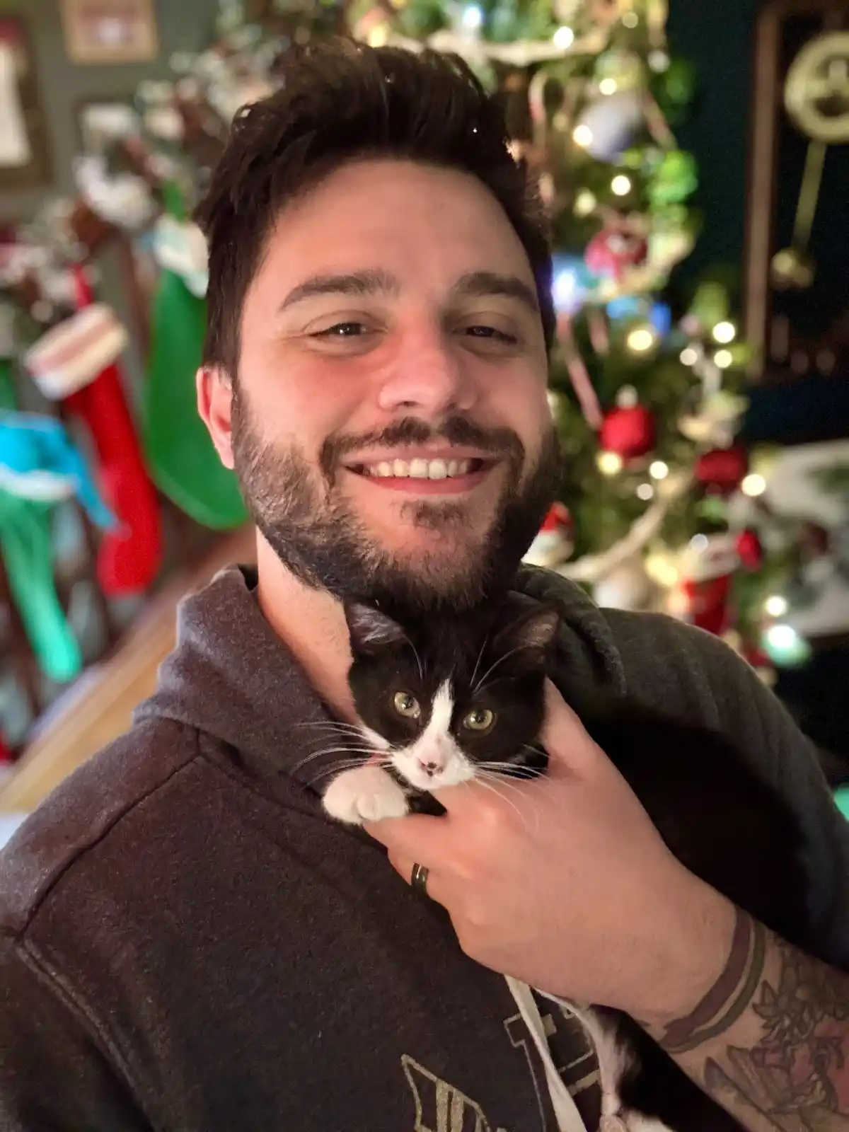 A man with a beard and dark hair, wearing a grey hoodie, smiles while holding a small black-and-white cat against his chest…
