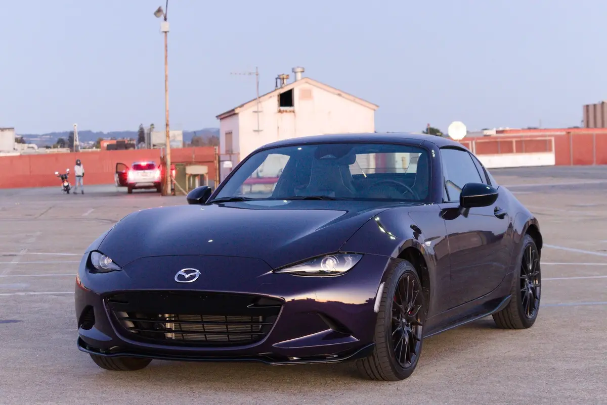 A dark purple Mazda Miata sports car is parked in an empty lot, its glossy finish reflecting the ambient light. In the…