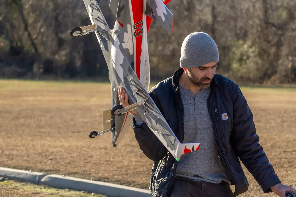 A man in a grey beanie and black jacket holds a large, detailed remote-controlled fighter jet model with red and grey…