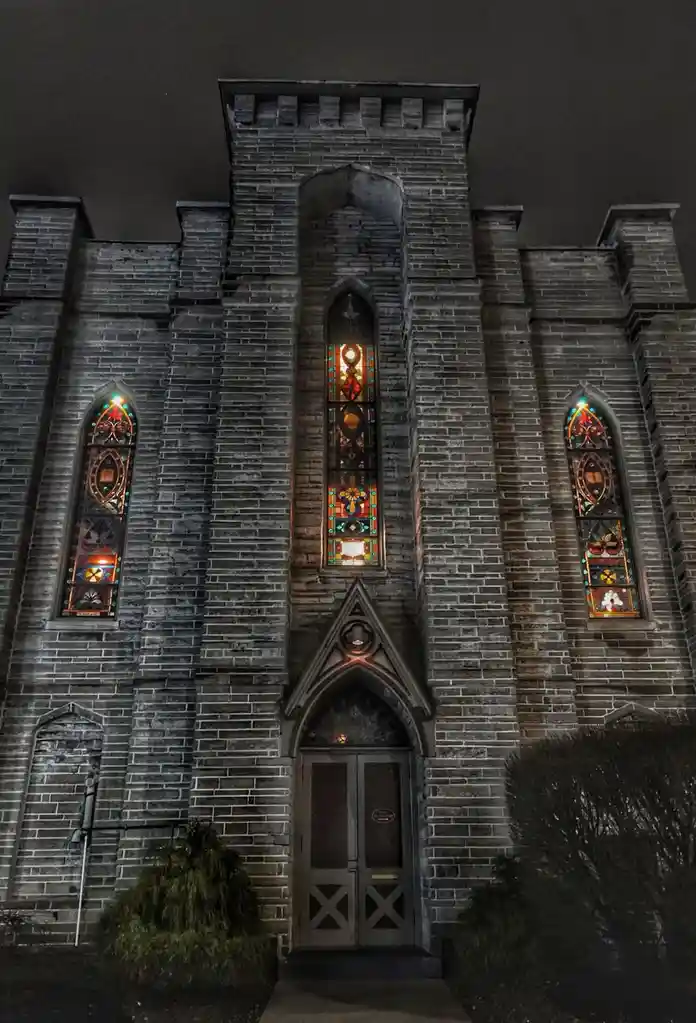 A nighttime photo of a Gothic-style brick church with three illuminated stained-glass windows and a central wooden door. The…