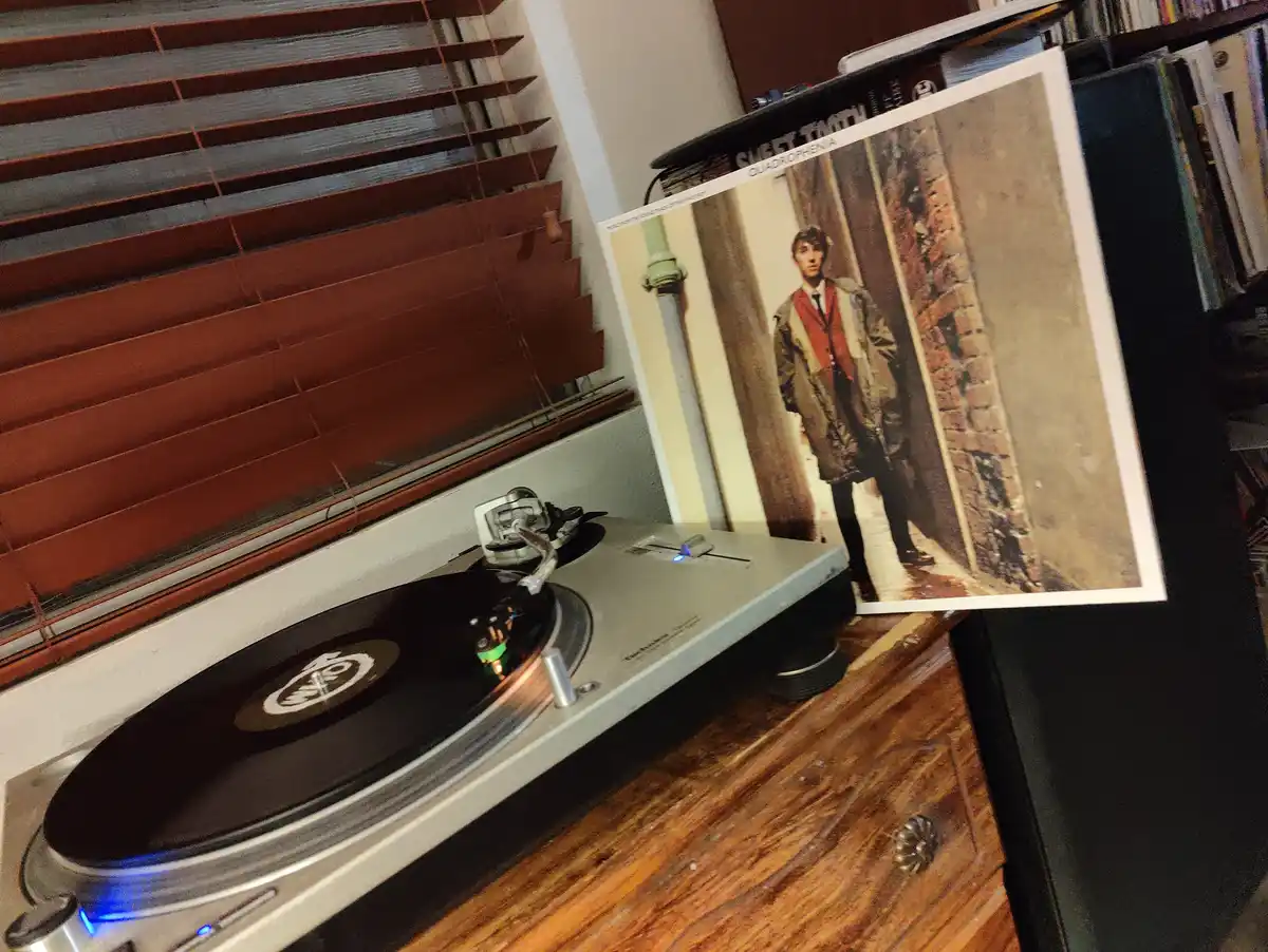 A vinyl record player sits on a wooden surface with a record spinning, next to the album cover of 'Quadrophenia' by The Who…