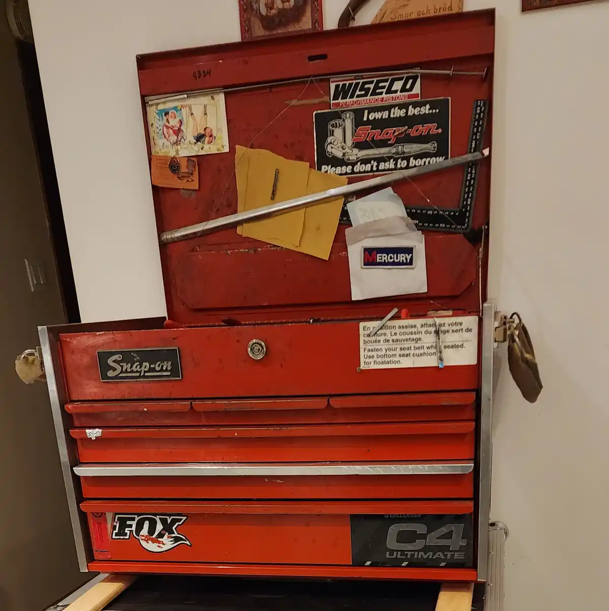 A red Snap-on tool chest with its top lid open, covered in stickers and memorabilia including a 'WISECO' sign, a 'MERCURY'…