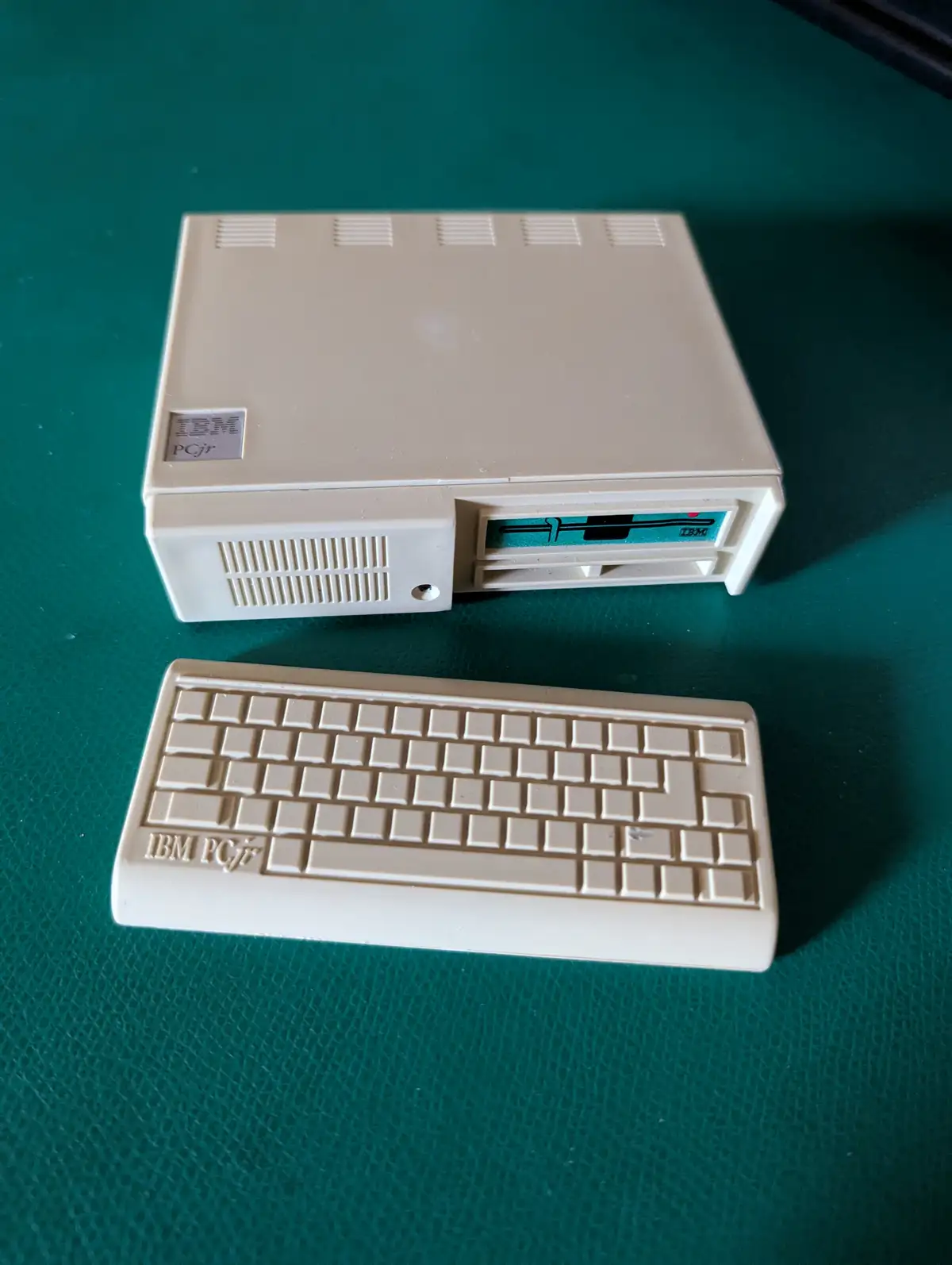 A beige plastic toy replica of an IBM PCjr computer and keyboard sits on a green textured surface. The toy computer and…