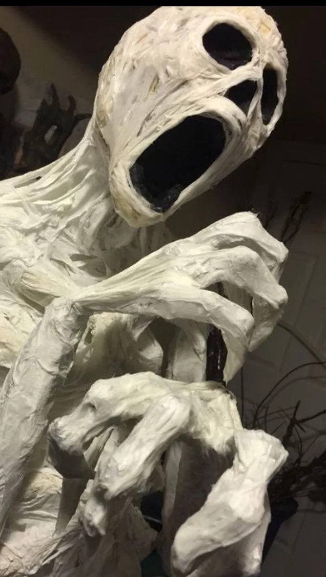 A close-up photo of a handmade, white, papier-mâché ghost decoration with a screaming face and claw-like hands, set against…