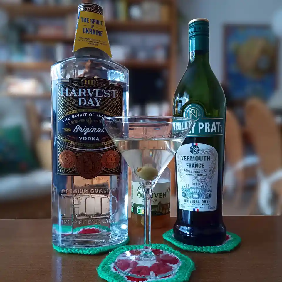 A still life photo shows a bottle of Harvest Day vodka, a bottle of Noilly Prat vermouth, and a martini glass with an olive…